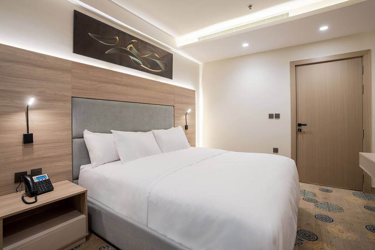 A modern hotel room with a large bed, a bedside telephone, a wall-mounted artwork, and a wooden door.