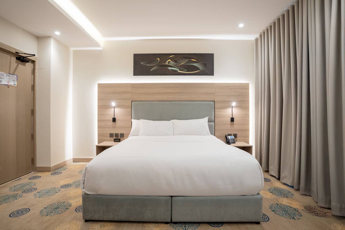 A modern hotel room with a large bed, wooden headboard, two bedside tables with lamps, and a large window with curtains.
