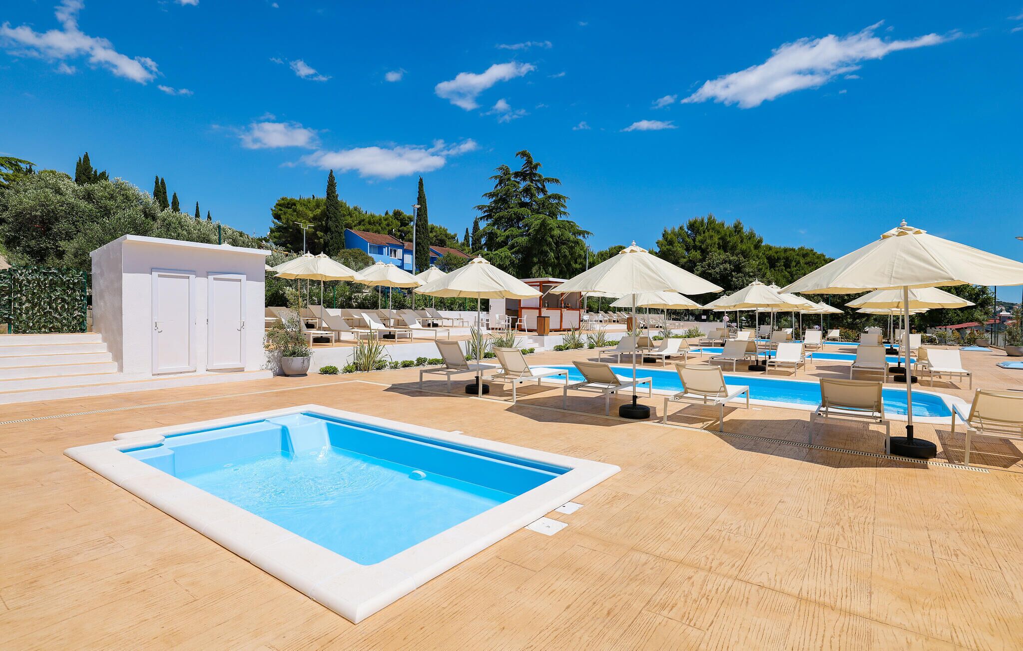 A pool area with multiple sun loungers and umbrellas, a small pool, and a white building with doors.