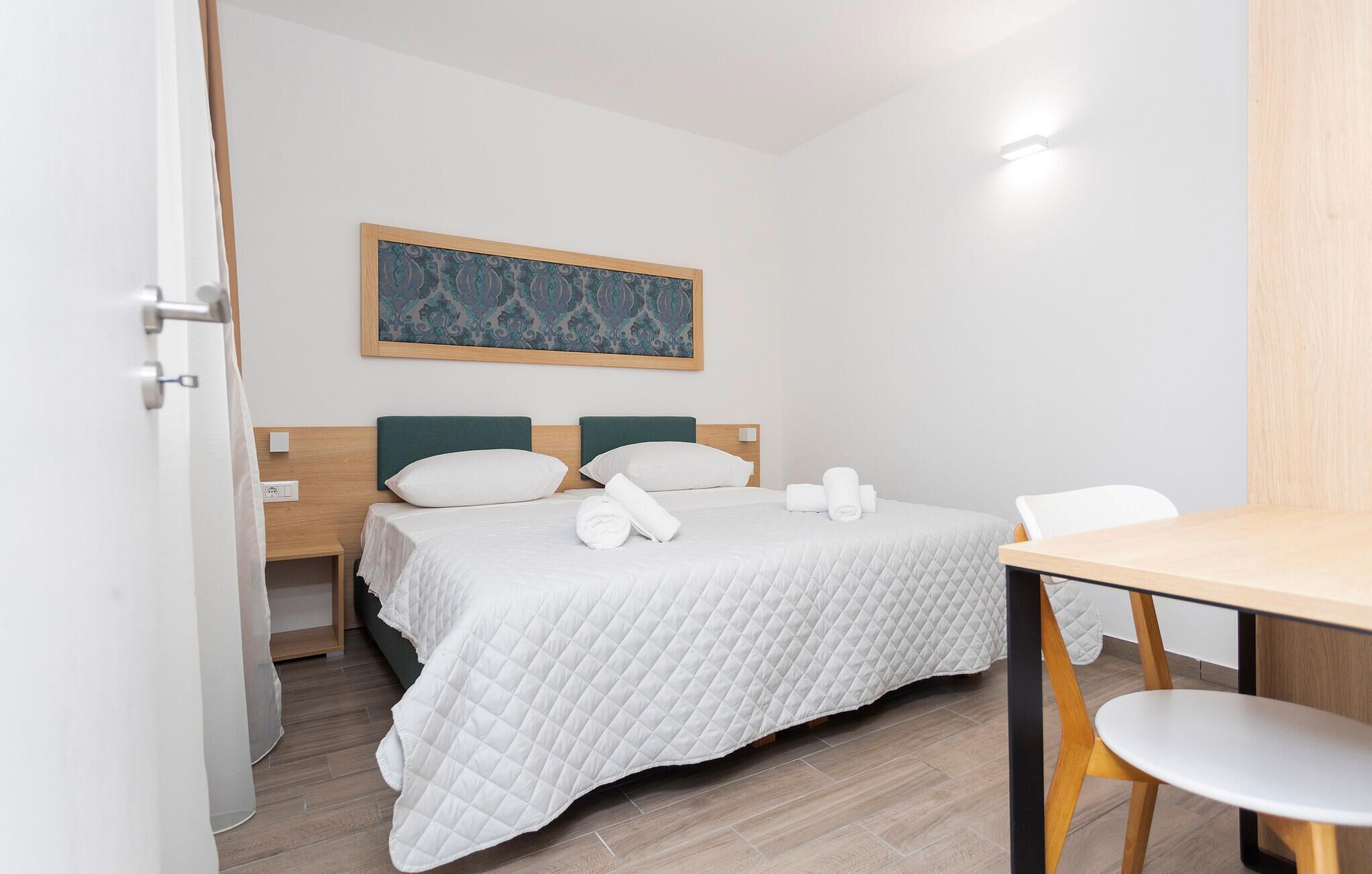 A hotel room with a bed, two pillows, a quilted blanket, a painting on the wall, a wooden nightstand, a white chair, and a table.