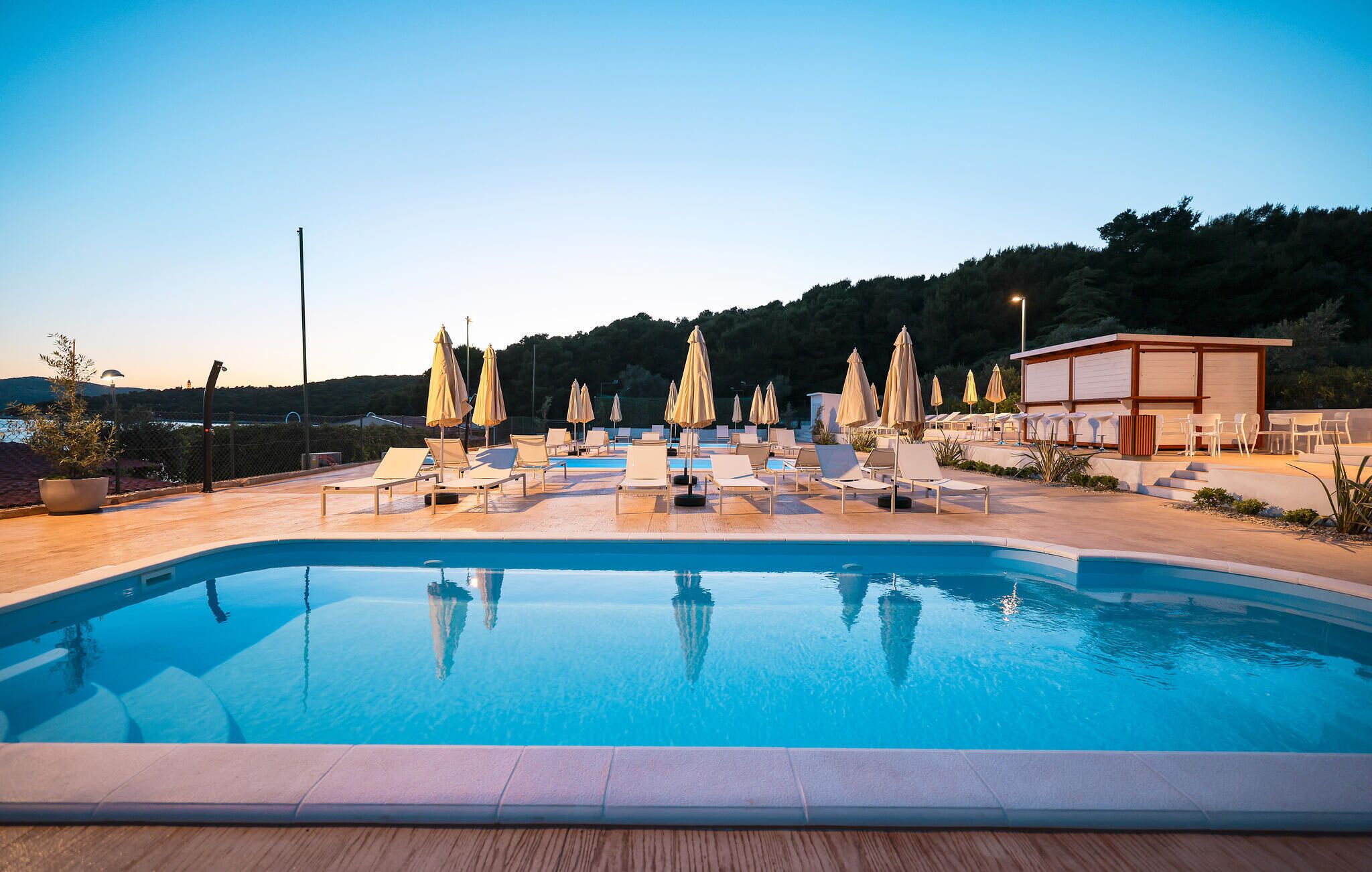 Une piscine entourée d’une terrasse en bois, agrémentée de chaises longues et de parasols, et bordée d’un bâtiment dont l’enseigne est illuminée.