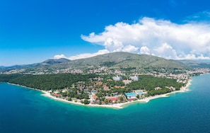 An aerial view of a coastal town with a mountain in the background, surrounded by clear blue waters and a small peninsula with buildings.