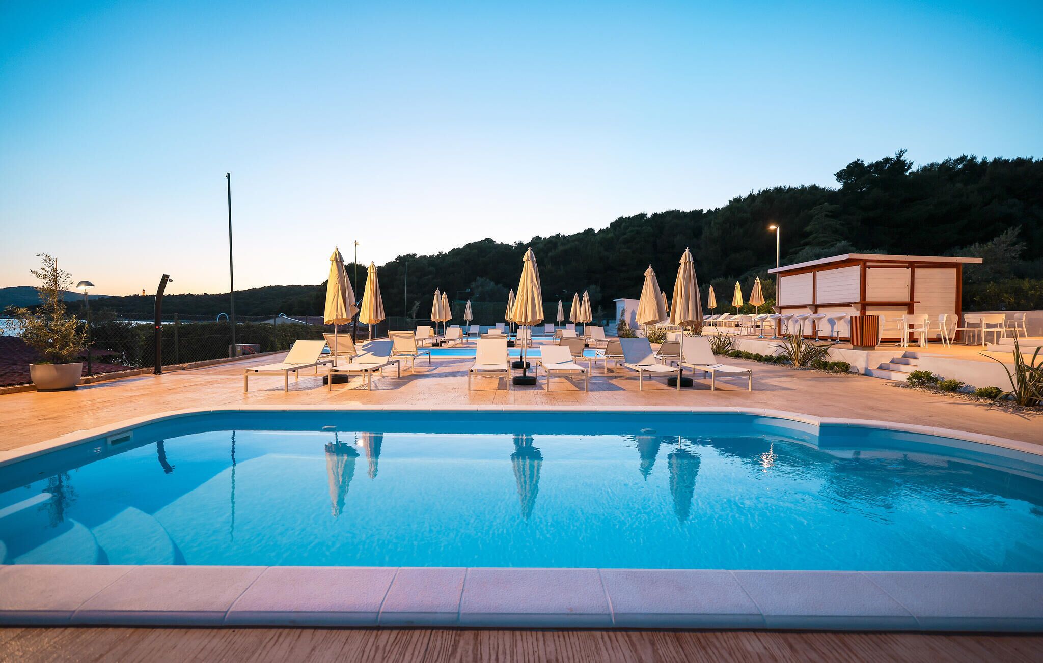 A swimming pool with lounge chairs and umbrellas, surrounded by a wooden deck and a building with a lighted exterior.
