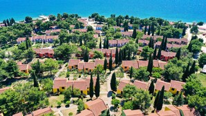 An aerial view of a coastal residential area with houses arranged in rows, surrounded by greenery and trees, with a clear view of the sea.