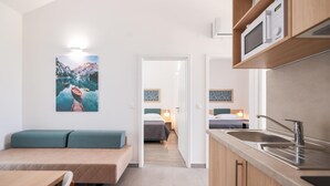 A compact hotel room with a kitchenette, a bed, a seating area, and a painting of a mountain lake.