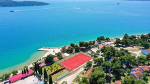 An aerial view of a coastal area with a clear blue sea, a small island, and a sports complex with a red court.