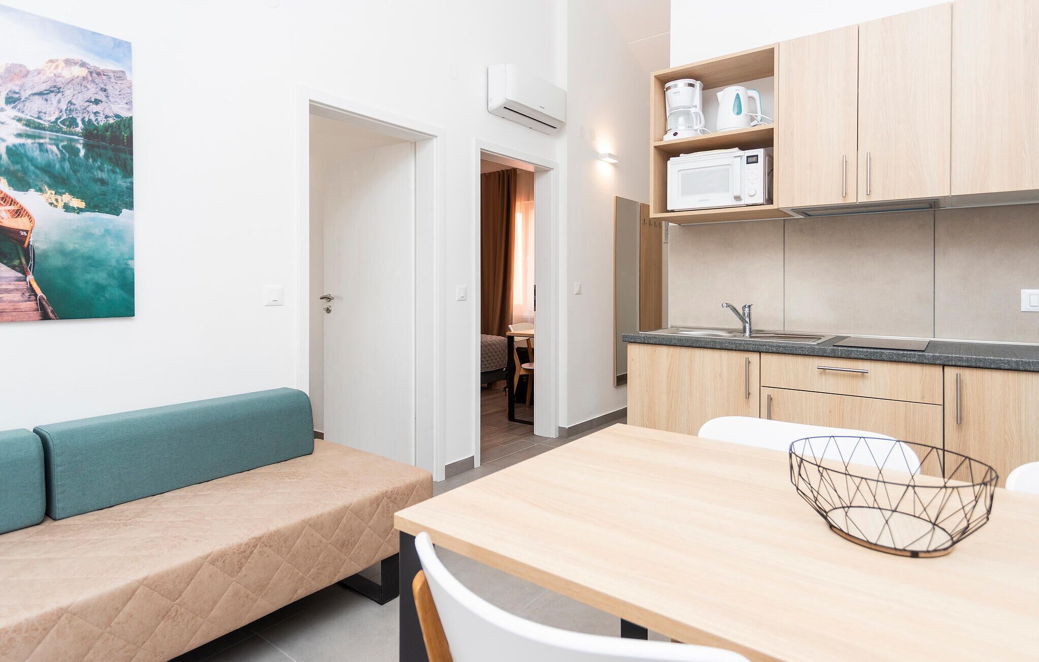 Une cuisine moderne équipée d’un four à micro-ondes, d’une machine à café et d’une bouilloire sur le plan de travail. On y trouve une table à manger en bois, un panier en fil métallique et un banc avec un coussin. Un tableau représentant un paysage de montagne est accroché au mur.