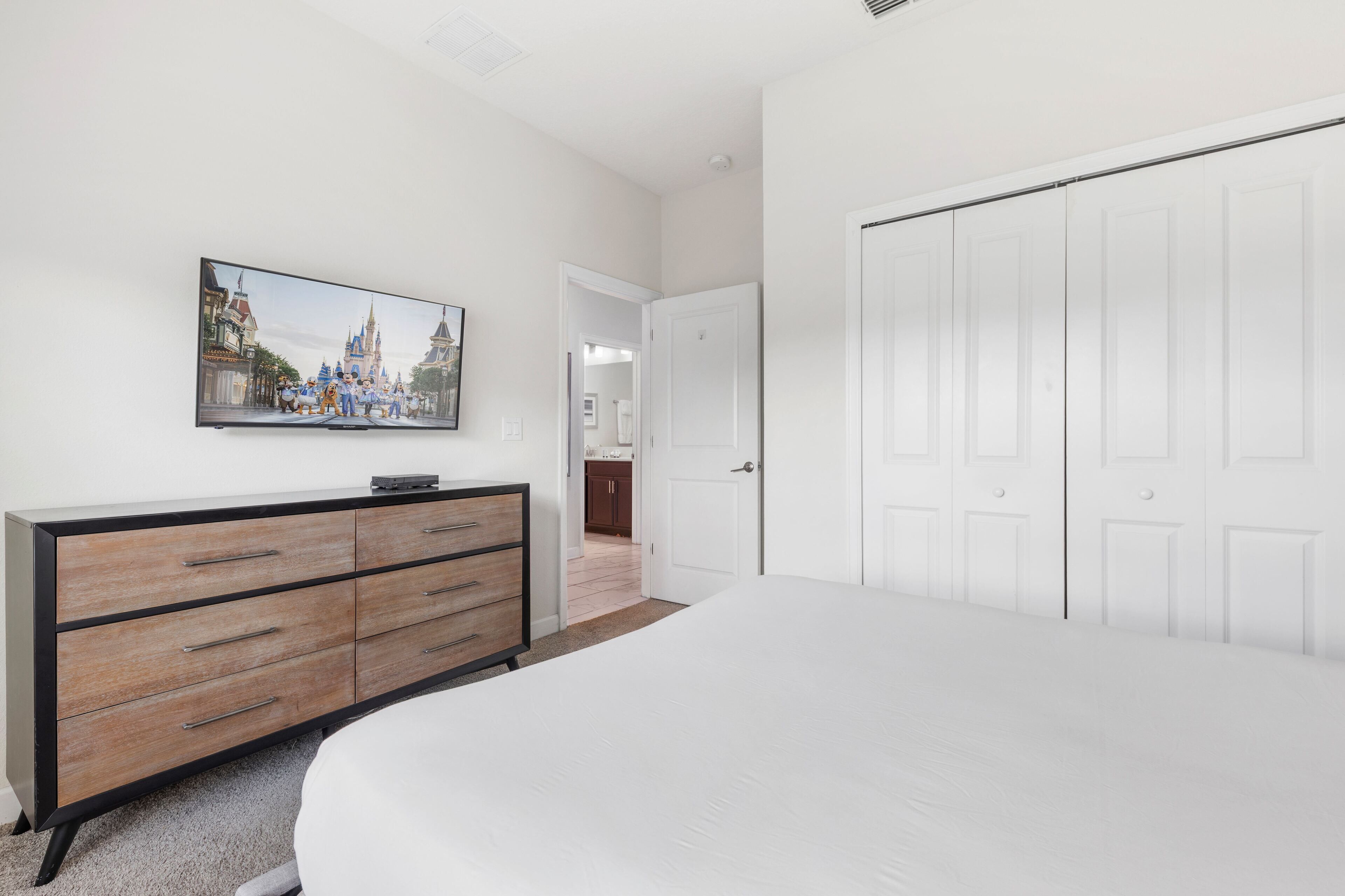 A bedroom with a large bed, a dresser, a television mounted on the wall, and a closet with multiple doors.