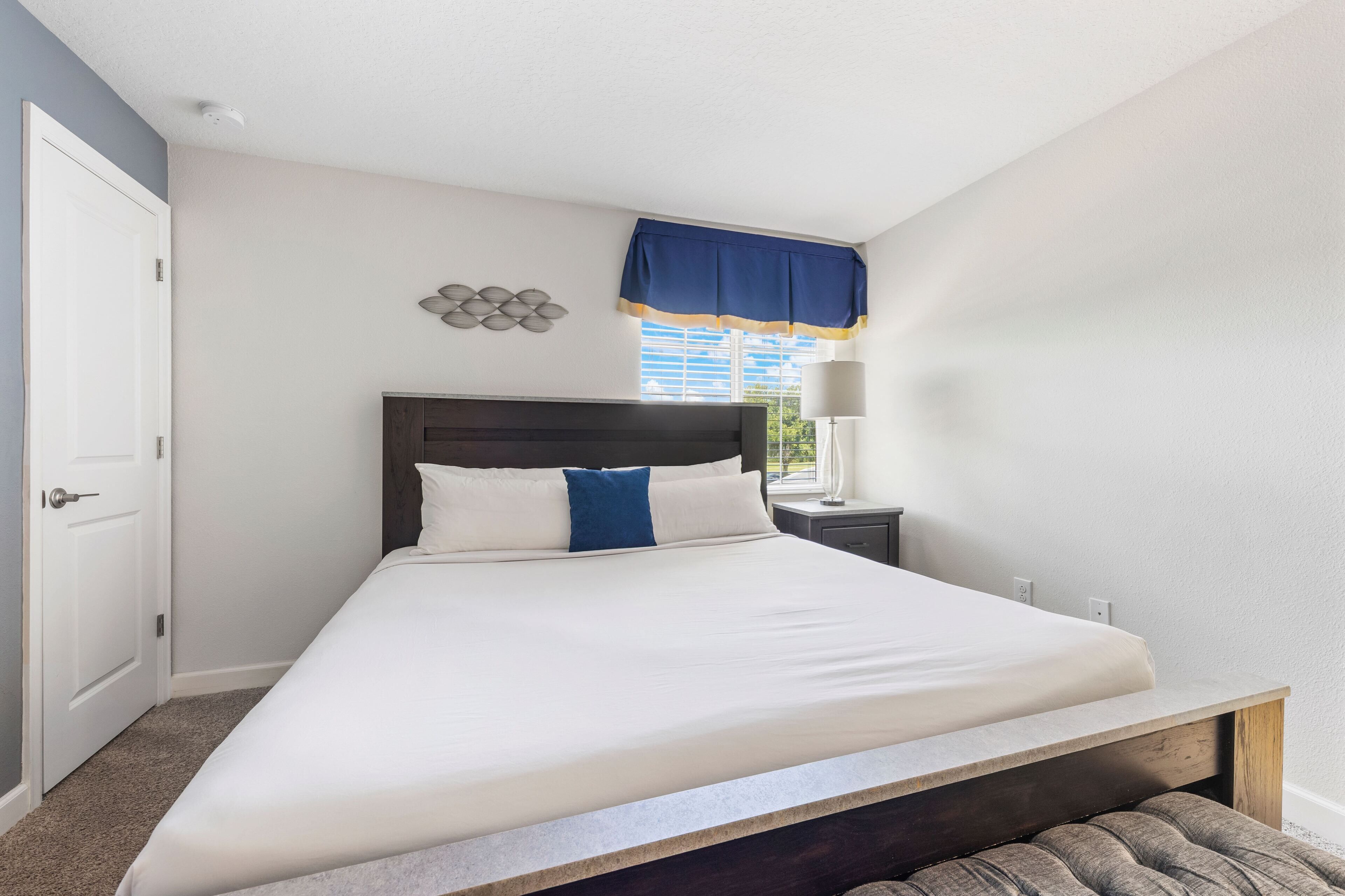 A neatly made bed with a headboard, a bedside lamp, and a window with a blue valance.