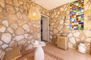 A stone-walled room with a wicker chair, a white table with teapots, a wooden side table, and a colorful stained glass window.
