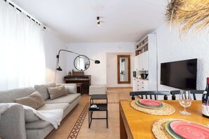 Una sala de estar con sofá, una mesa de centro, una mesa de comedor con individuales, televisor y una cocina al fondo.