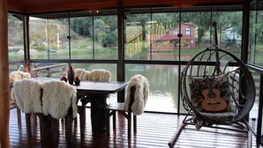 Eine Holzterrasse mit Tisch und Stühlen, einem Hängestuhl und einer Gitarre, sowie Blick auf ein Haus und Bäume durch große Fenster.