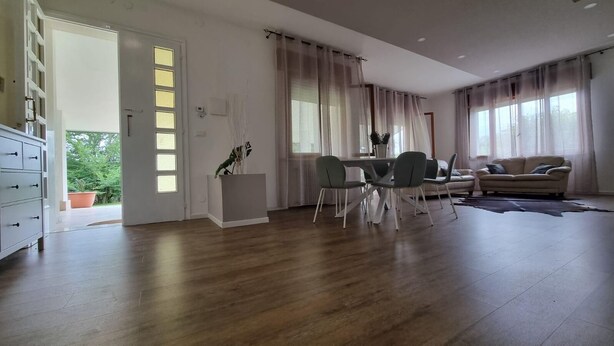 A spacious living room with a wooden floor, a dining area with a table and chairs, a sofa, and a large window with curtains. - Villa Savalin, a refined residence nestled in the quiet of Taglio di Po (Taglio di Po)