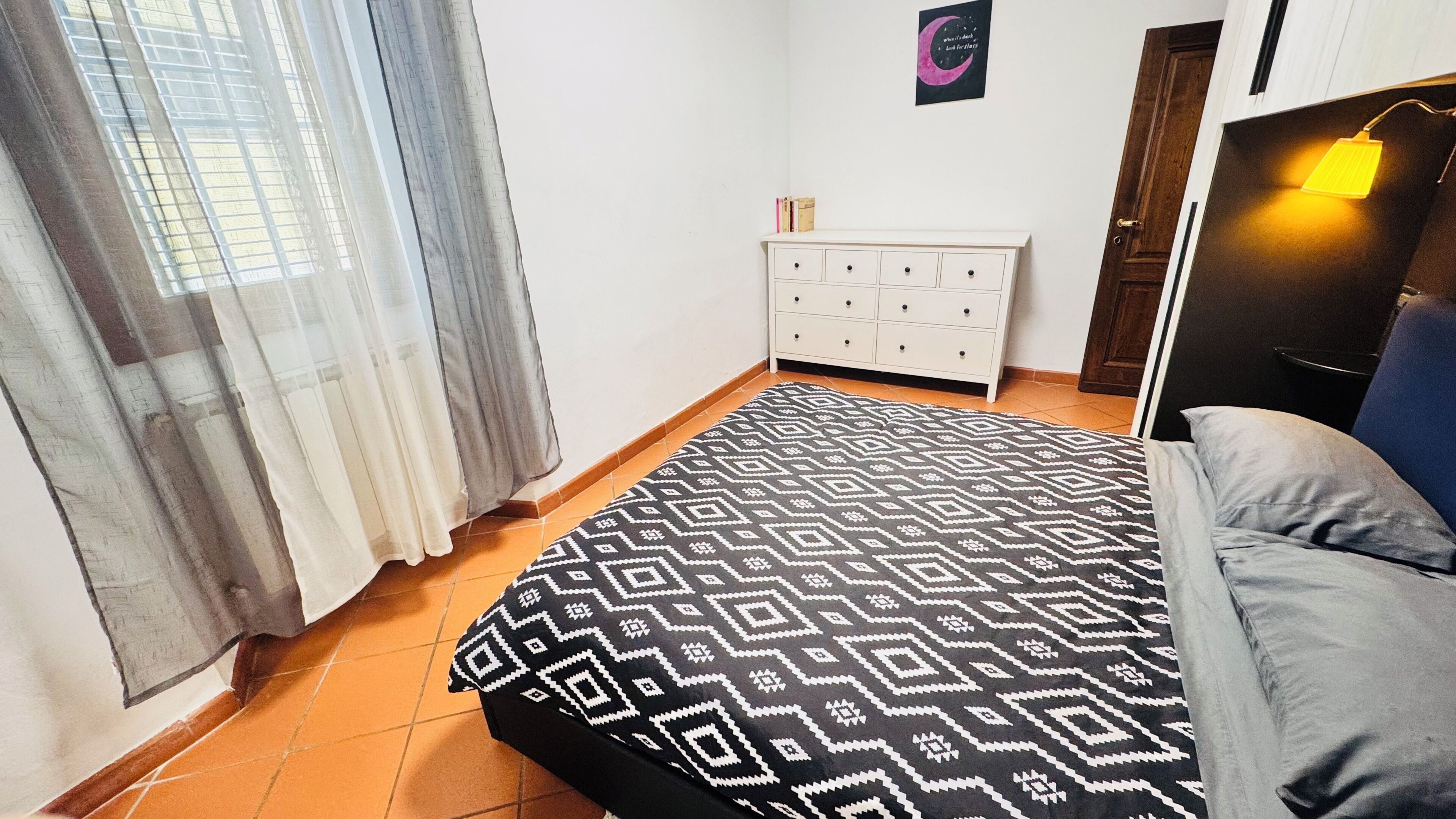 Een slaapkamer met een bed met een patroondiscreet, een witte dressoir, een deur met een lamp en een muur met een poster.
