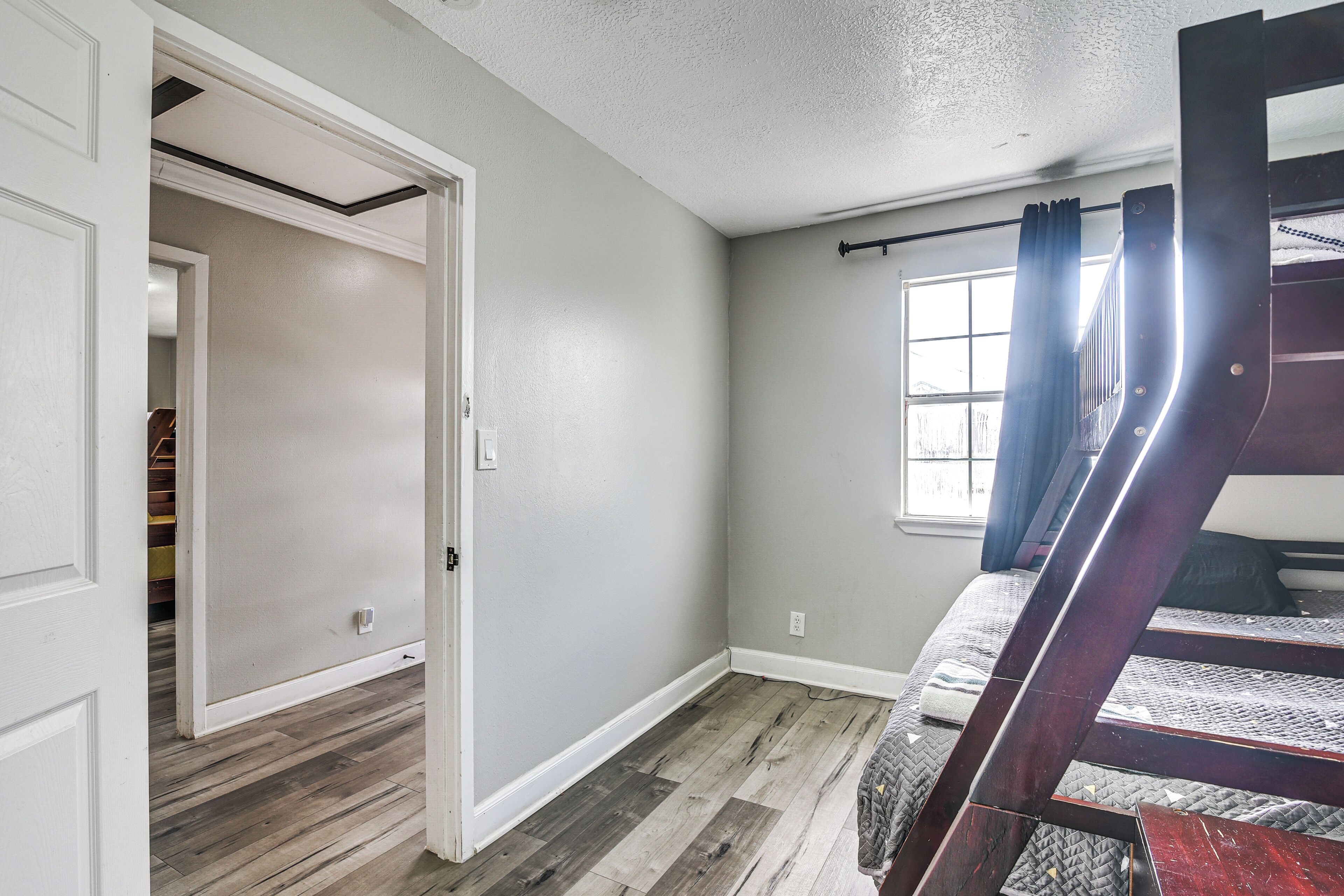 A bedroom with a bunk bed, a window with curtains, a wooden floor, and a door leading to another room.