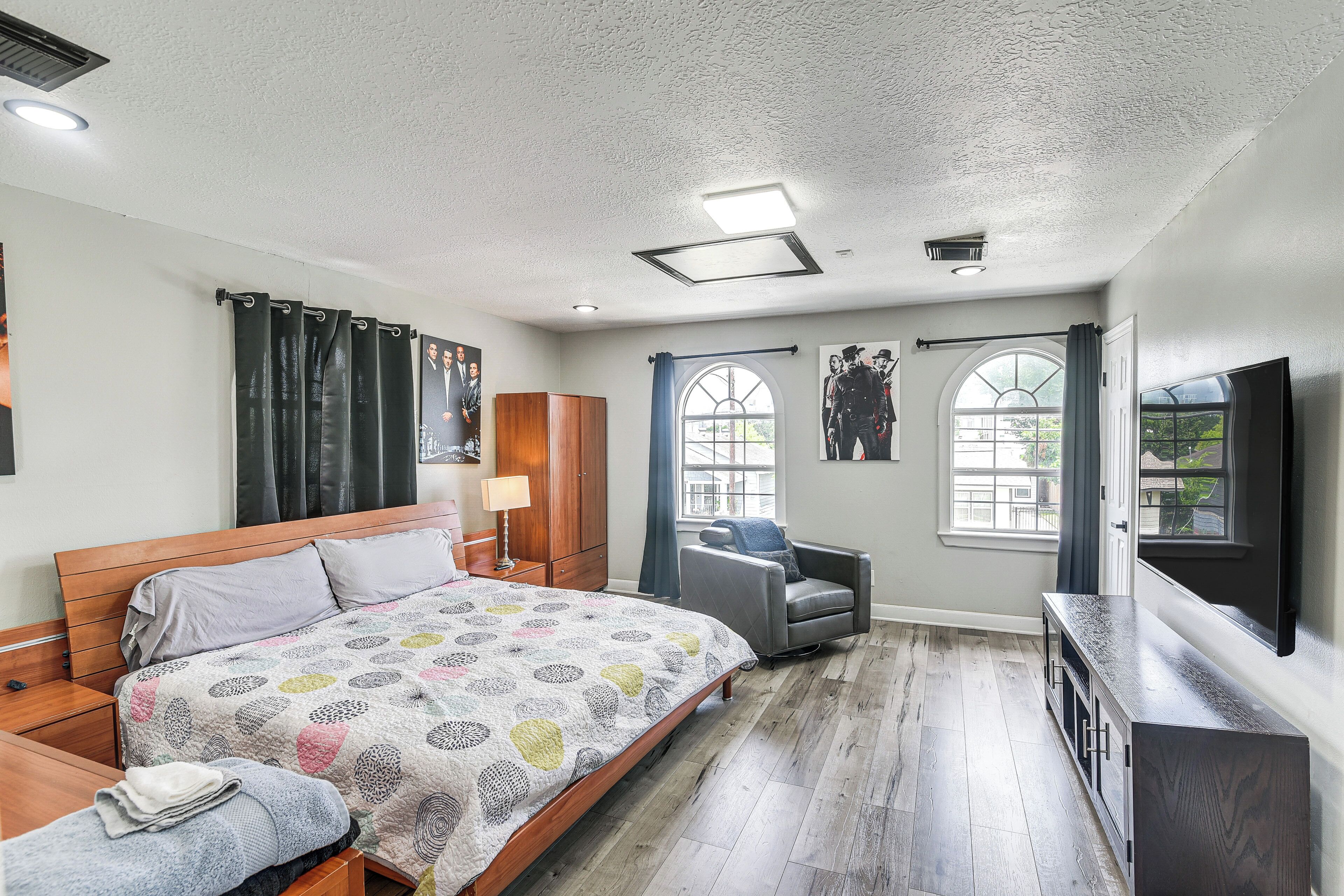 A bedroom with a wooden bed, a grey armchair, a TV, and a window with curtains.