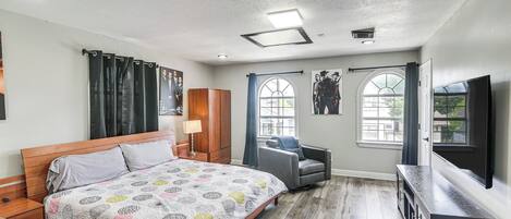 A bedroom with a wooden bed, a grey armchair, a TV, and a window with curtains.