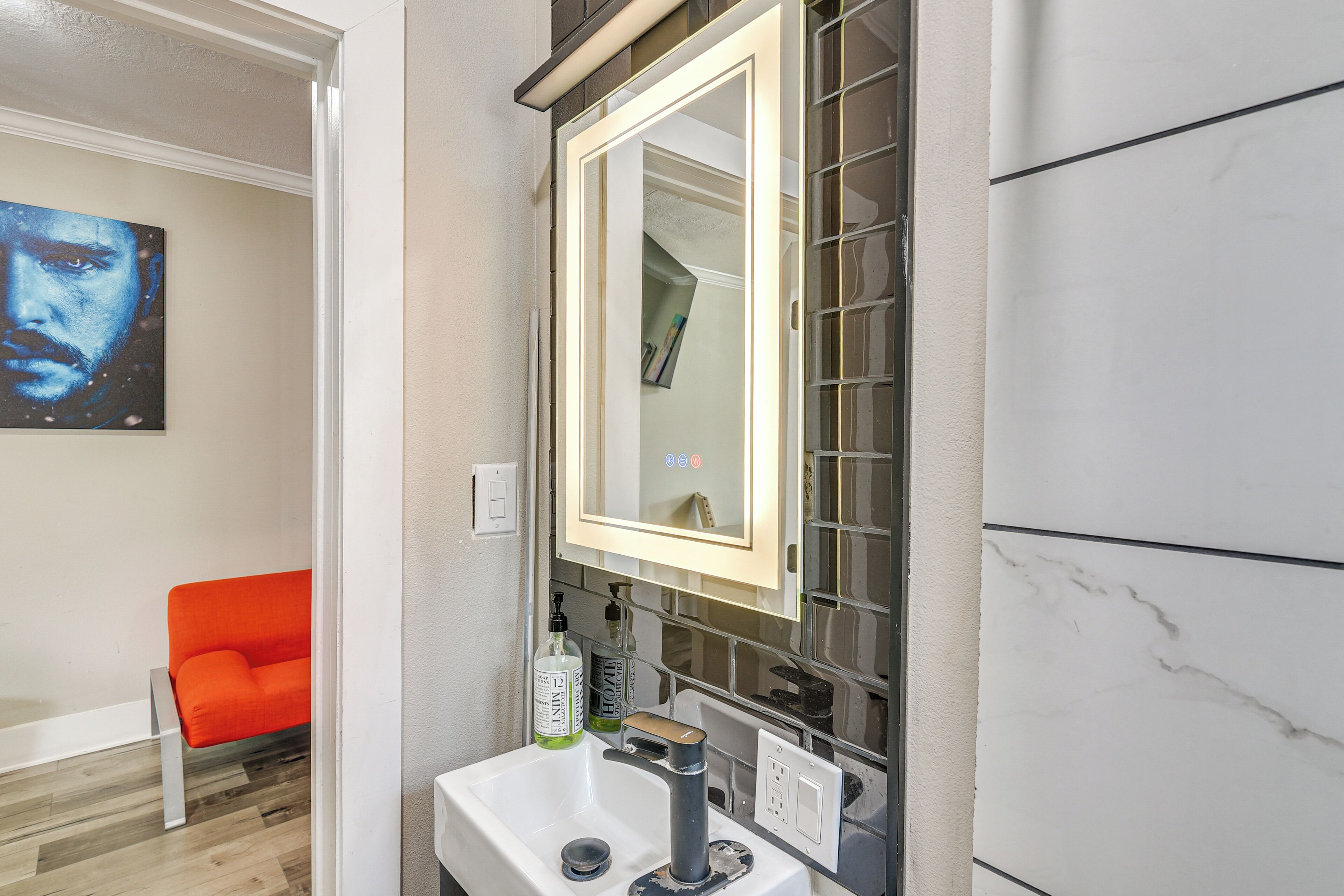 A bathroom with a white sink, a mirror with a light, and a red chair in the background.