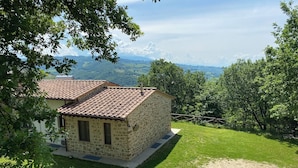 Una casa de piedra con techo de tejas, rodeada de césped y árboles, con vistas a colinas distantes.