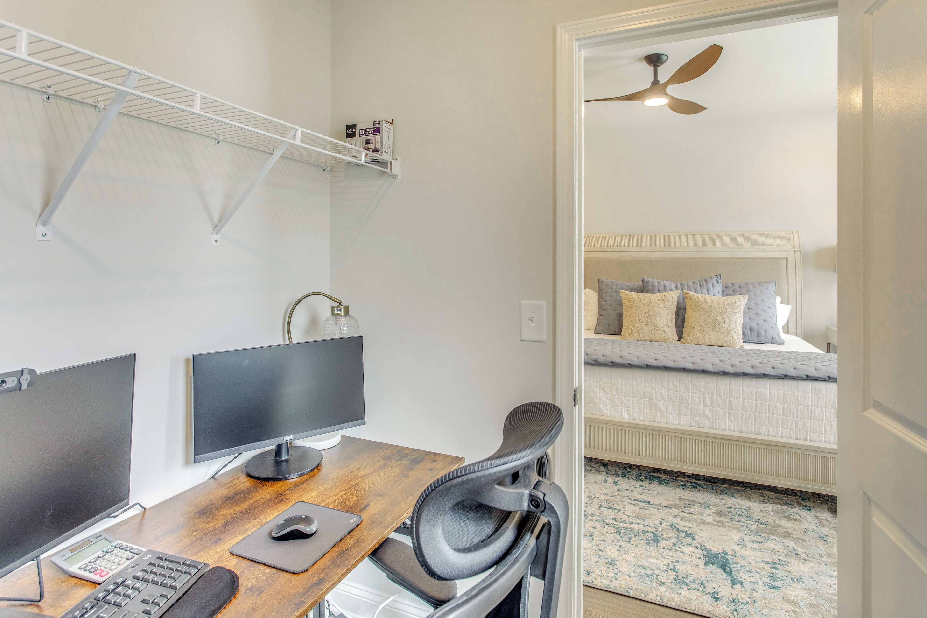 A room with a wooden desk, two monitors, a chair, a bed with pillows, a ceiling fan, and a shelf with items.
