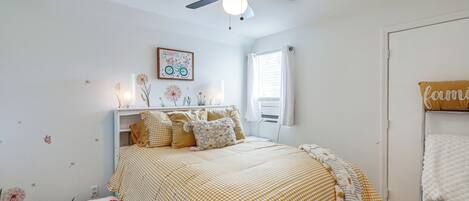 Un dormitorio con una cama, ventilador de techo, ventana y estante en la pared con objetos decorativos.