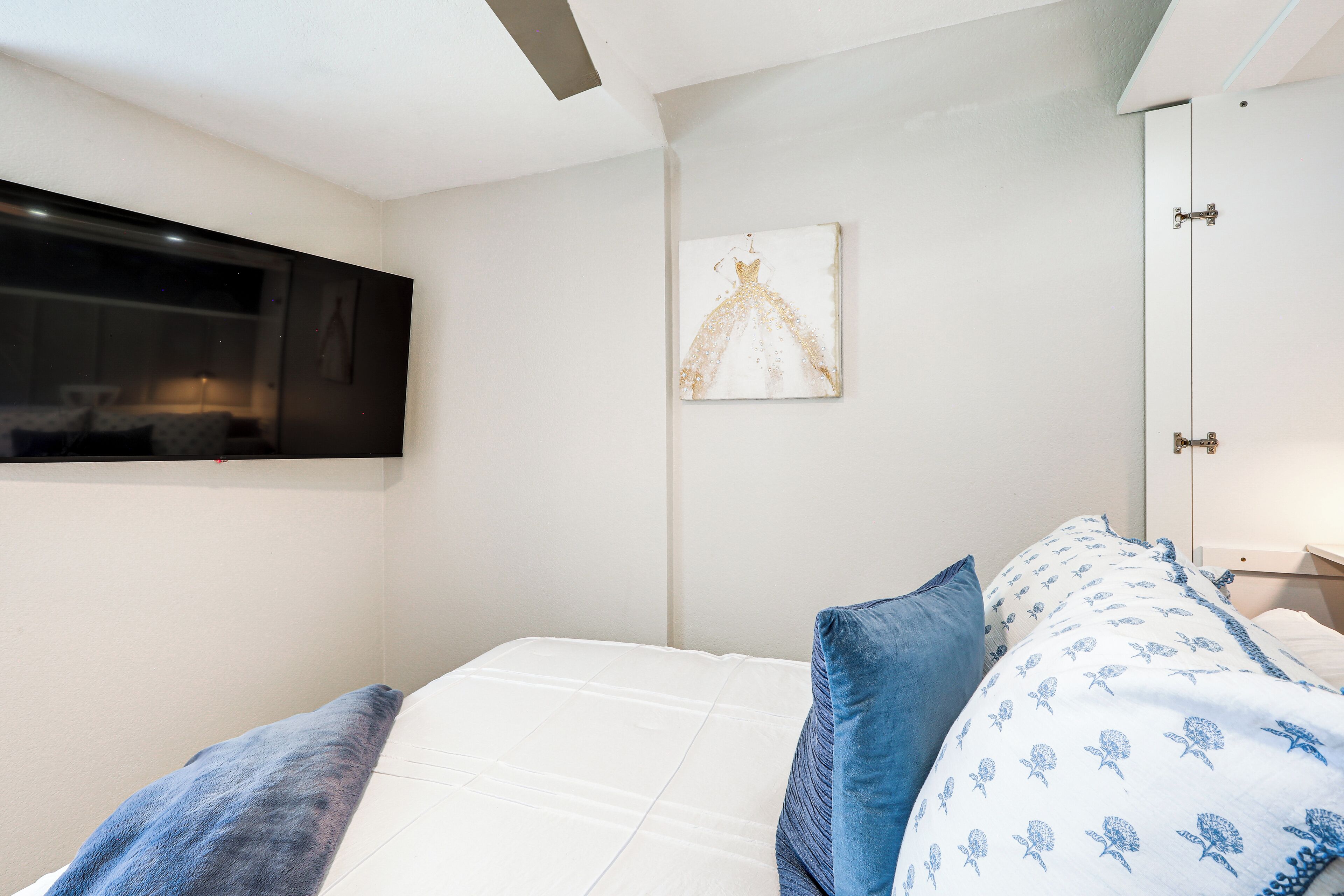 Un dormitorio con cama, almohadas, un televisor montado en la pared y un cuadro.