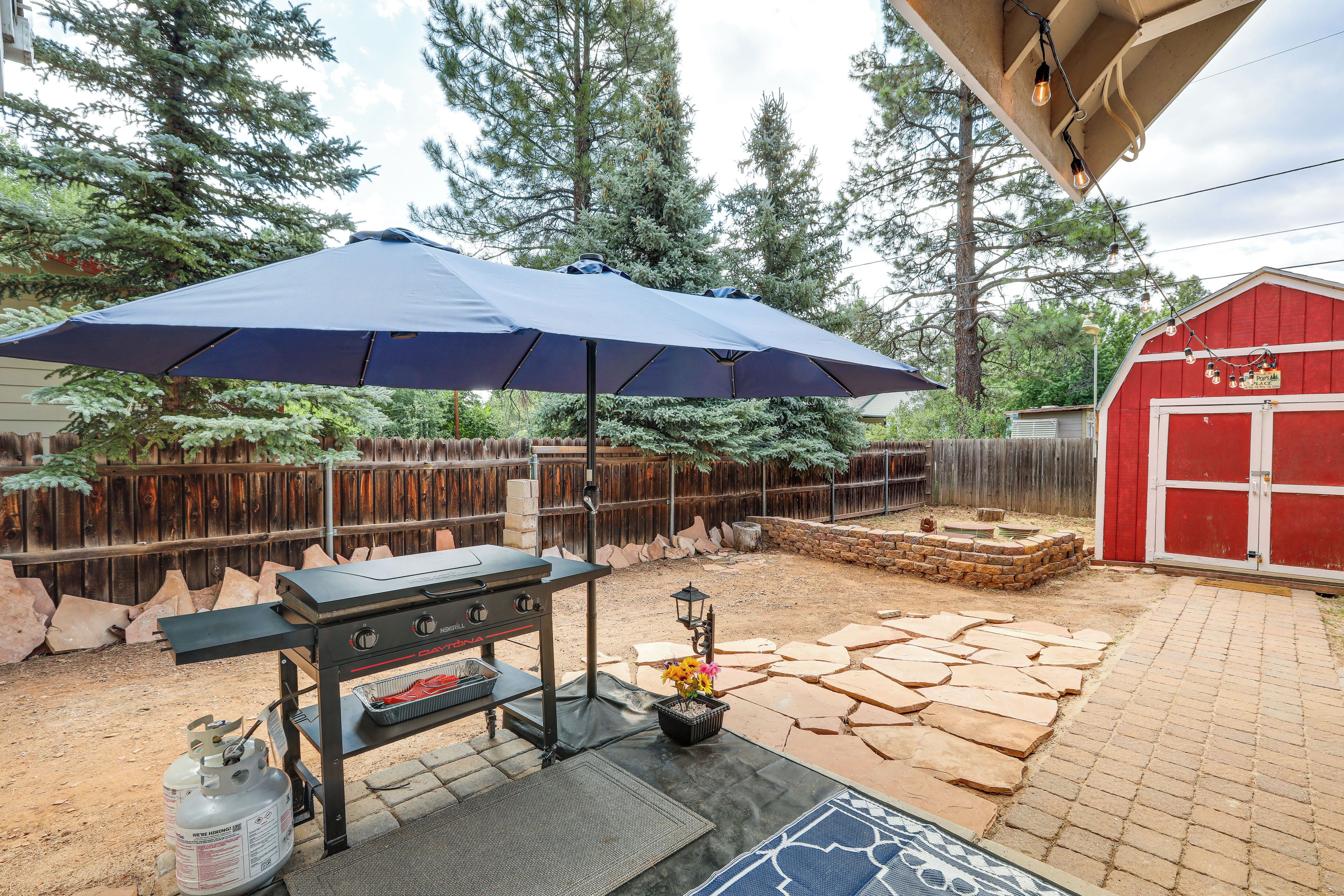 Un jardin avec un barbecue, un parasol, une bouteille de gaz et un abri de jardin rouge.