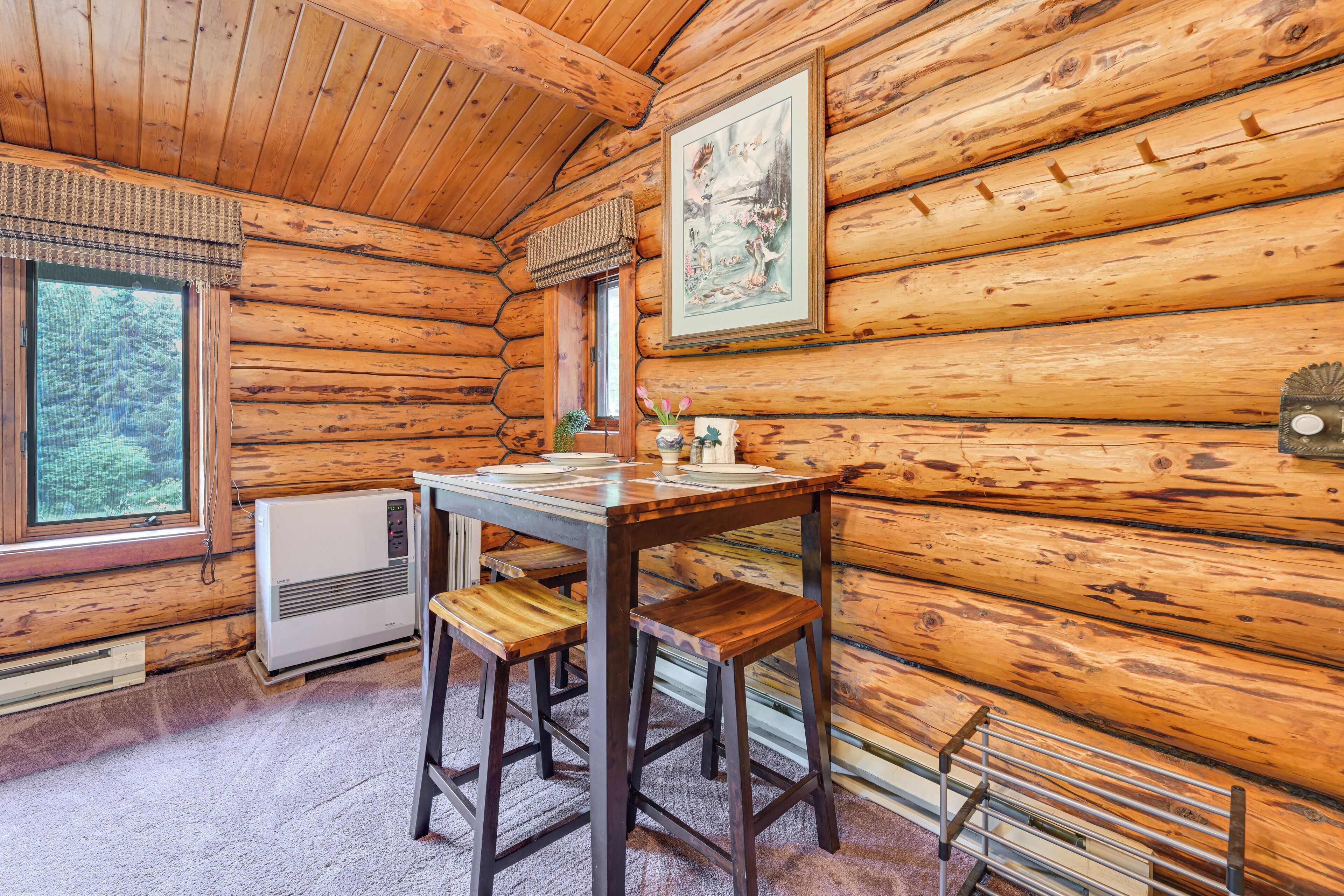 Ein Holzblockhaus-Interieur mit einem Holztisch, zwei Hocker, einer Heizung, einem Fenster und einem gerahmten Bild an der Wand.