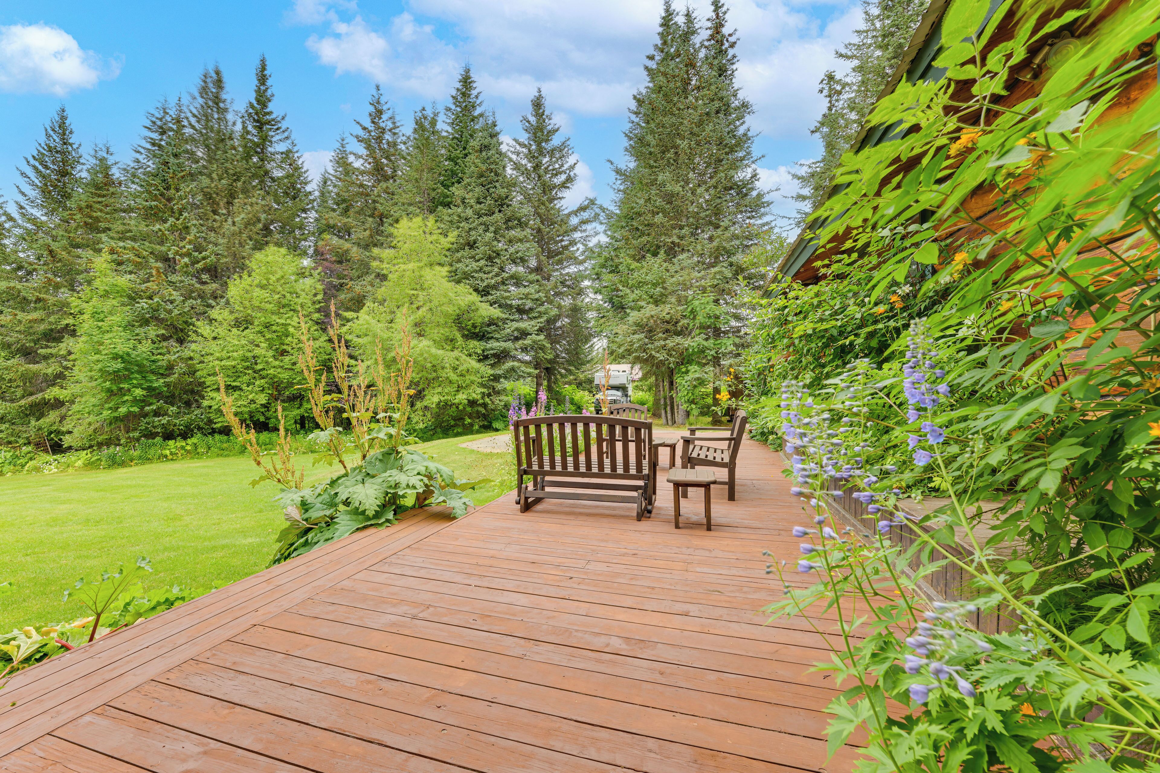 Una terraza de madera con un banco y sillas, rodeada de un jardín exuberante y árboles altos.