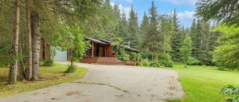 A log cabin with a concrete path leading to the entrance, surrounded by dense forest.