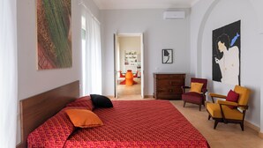 A bedroom with a large bed, two armchairs, a dresser, and a dining area in the background.
