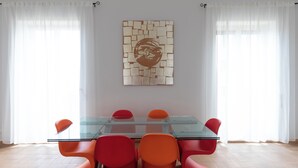 A modern dining area with a glass table and orange chairs, white curtains, and a mosaic artwork on the wall.