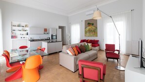 A modern living room with a glass coffee table, red and orange chairs, a beige sofa with colorful cushions, a red armchair, and a large window with a painting above it.