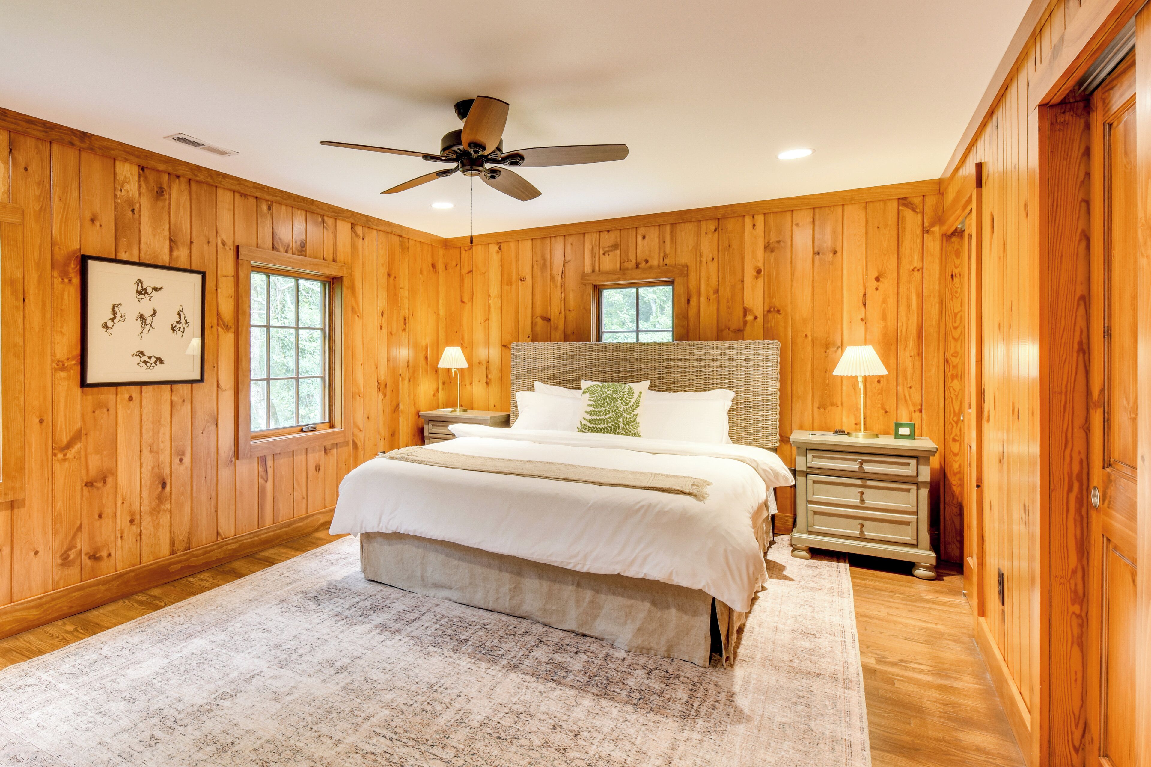 A bedroom with wooden walls, a bed with white bedding, a ceiling fan, two bedside tables with lamps, and a window.