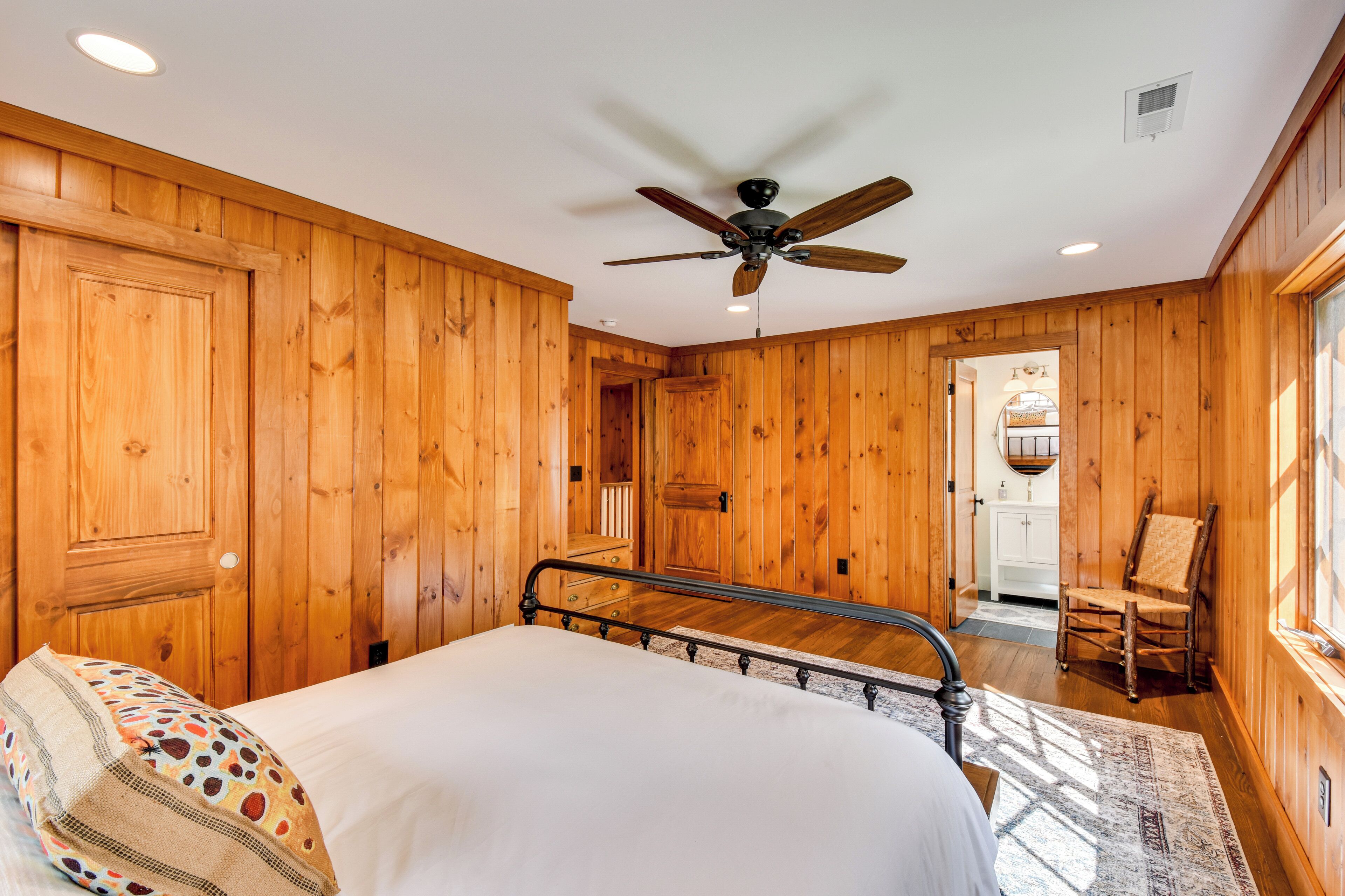 Une chambre avec des murs en bois, un ventilateur de plafond, un lit avec un coussin à motifs et une chaise.
