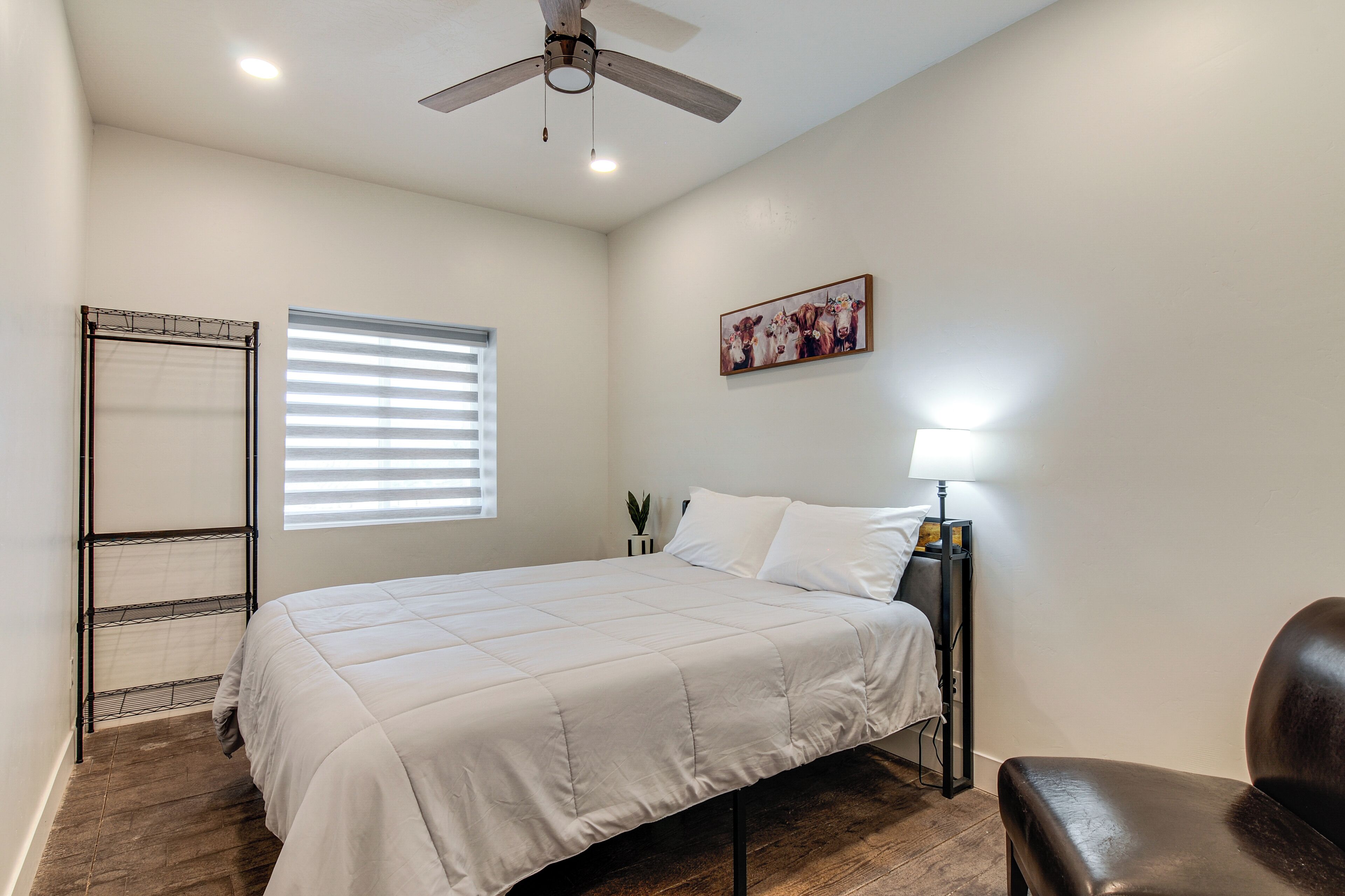 A bedroom with a bed, a ceiling fan, a window with blinds, a nightstand with a lamp, and a painting on the wall.