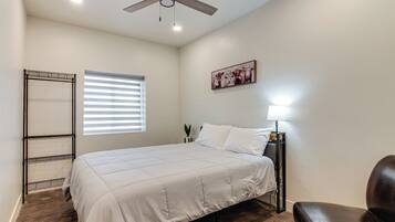 A bedroom with a bed, a ceiling fan, a window with blinds, a nightstand with a lamp, and a painting on the wall.