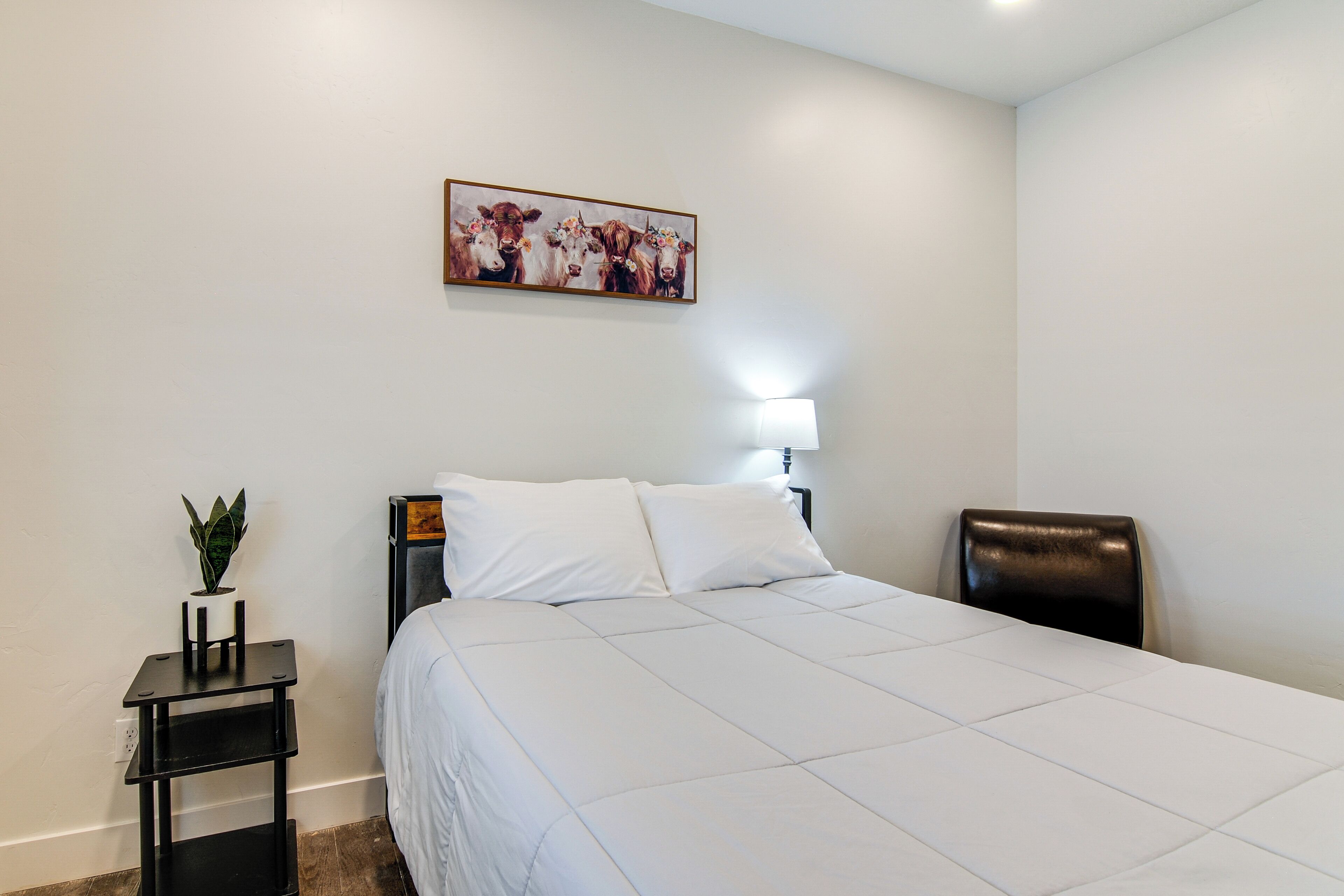 A hotel room with a bed, a nightstand with a plant, a lamp, and a painting on the wall.