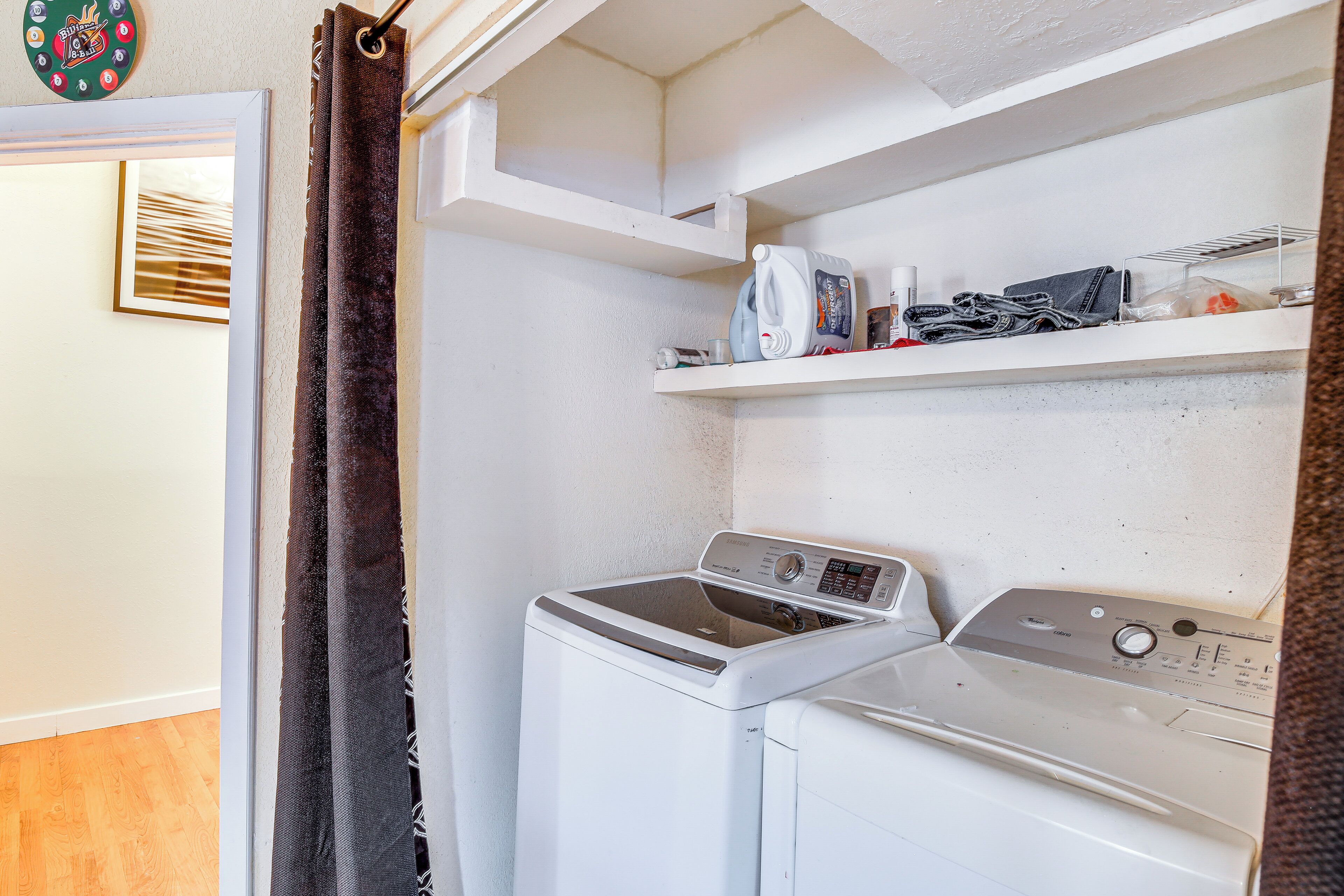 A laundry area with a washing machine and dryer, a shelf with cleaning supplies, and a door with a curtain.