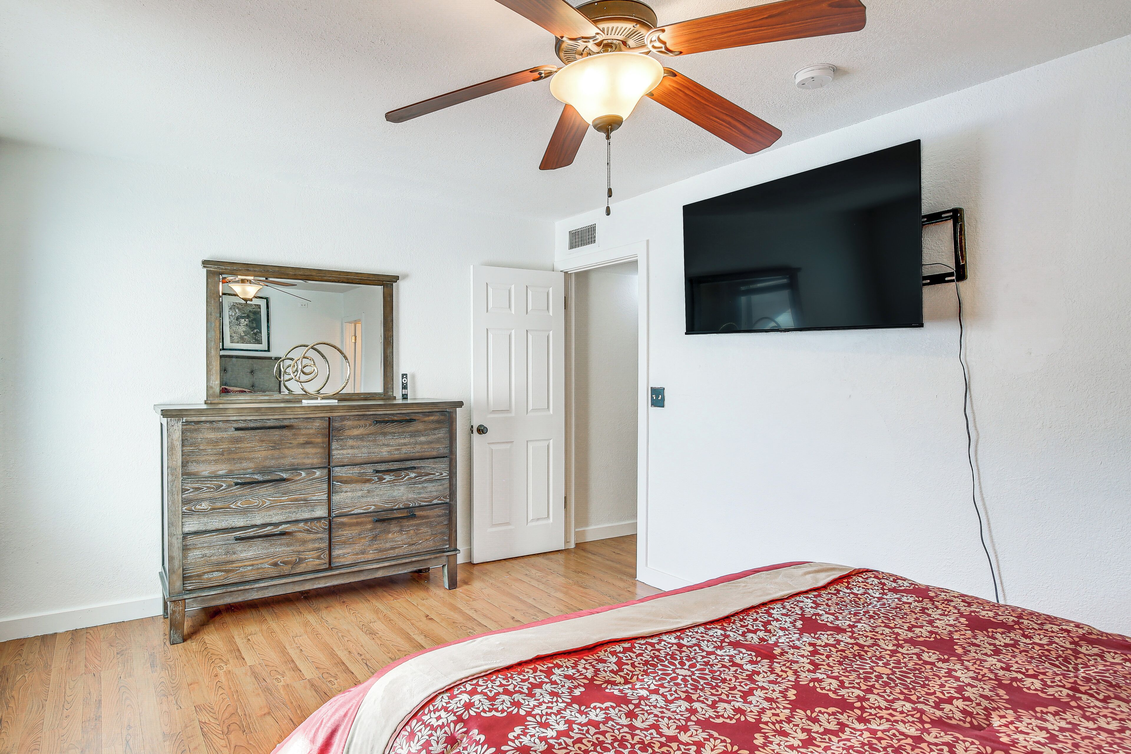 A bedroom with a wooden dresser, a ceiling fan, a bed with a red and white bedspread, and a mounted TV on the wall.