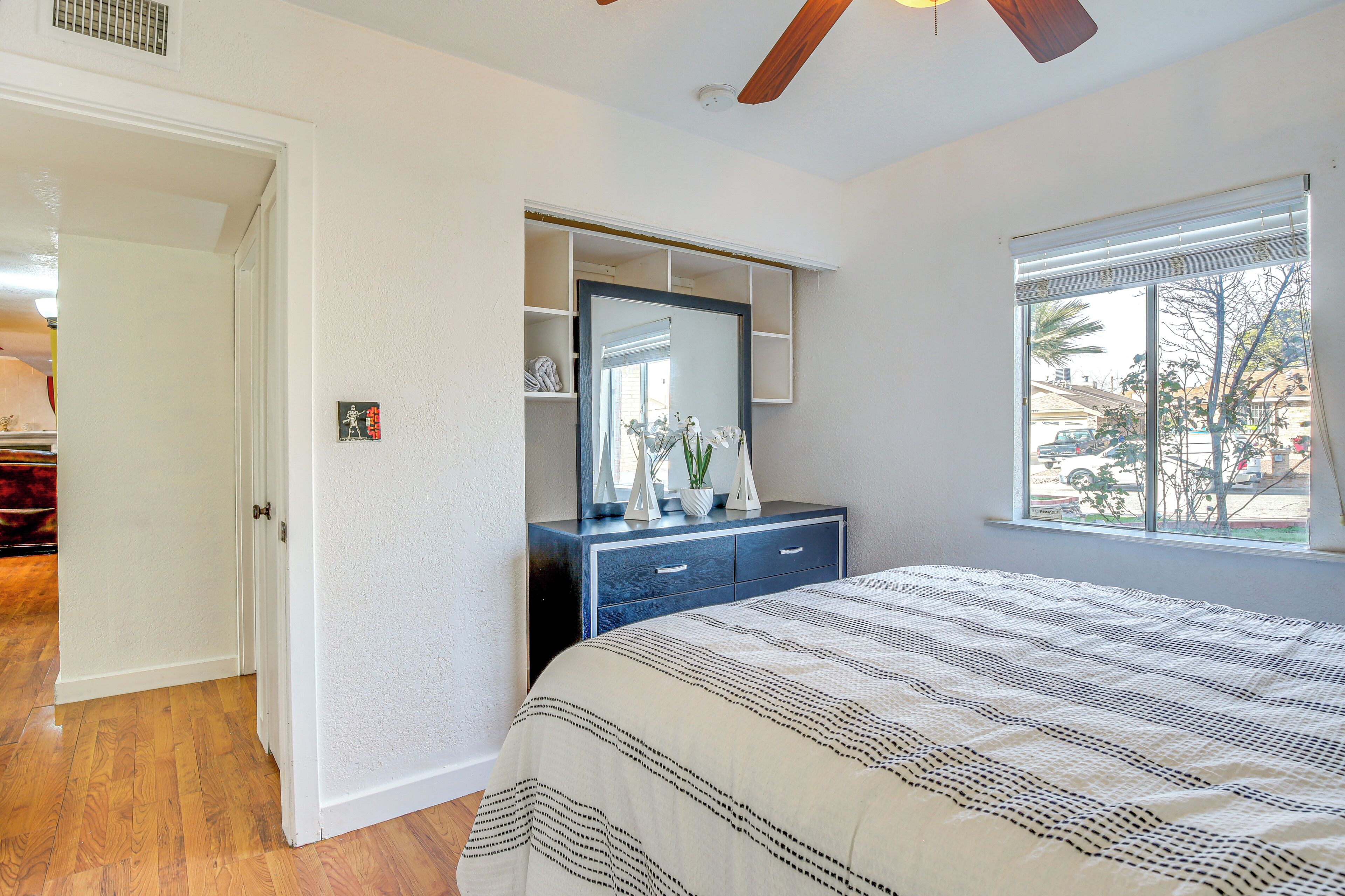 Un dormitorio con cama, cómoda con espejo, ventana con persianas y puerta que da a otra habitación.