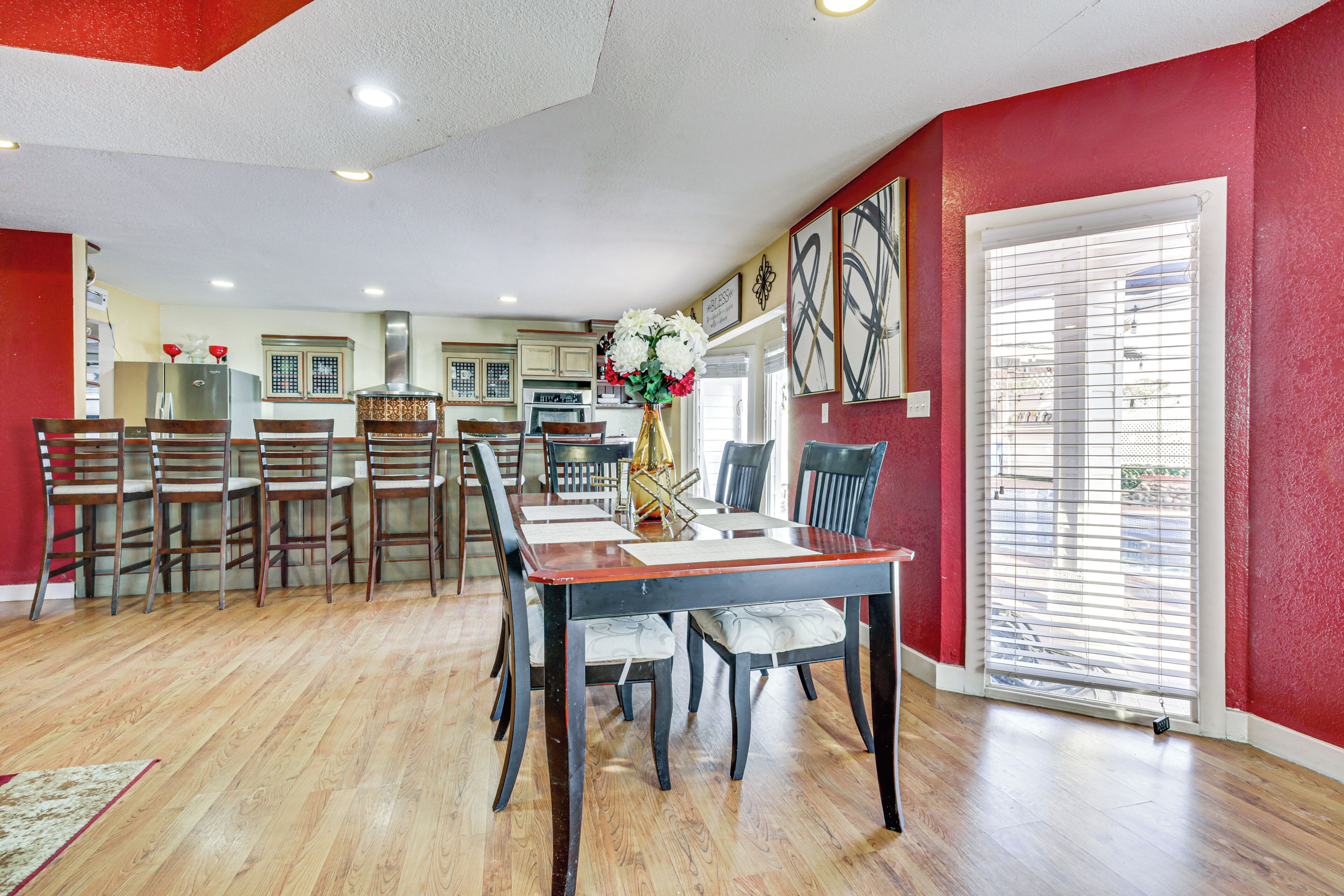 A dining area with a long table and chairs, a kitchen in the background, and a red wall with a large window.