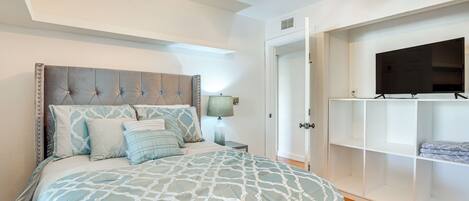 A bedroom with a tufted headboard, a bed with patterned bedding, a nightstand with a lamp, a wardrobe, and a ceiling fan.