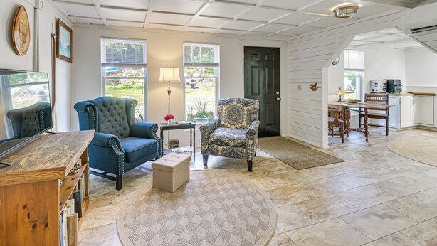 A spacious living room with a wooden coffee table, a blue sofa, and a patterned armchair. There is a dining area with a table and chairs, and a kitchen in the background. - Explore Coastal Maine! Cozy Apt w/ Yard & Garden (Orland)