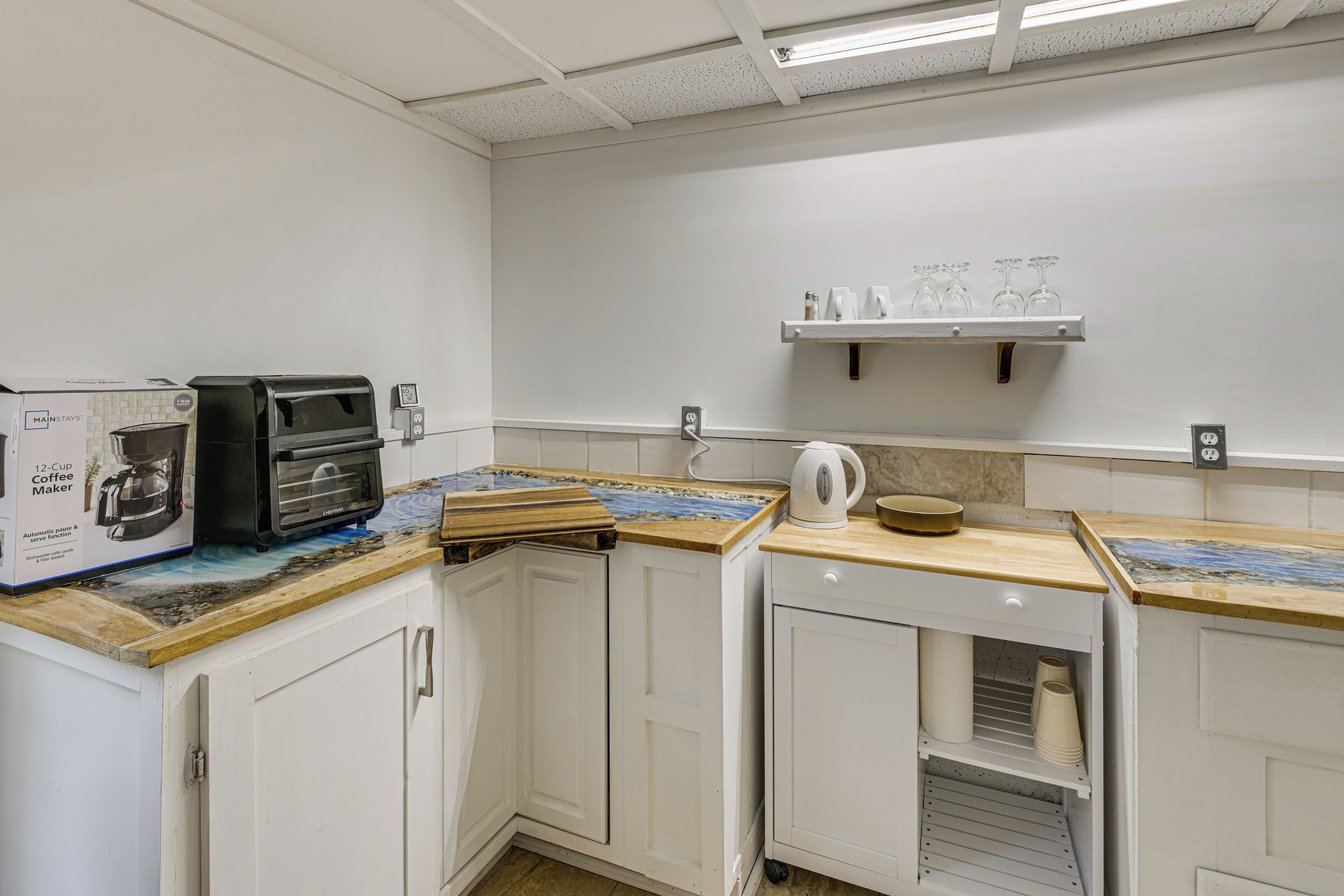 A kitchen with white cabinetry, a coffee maker, a kettle, and a shelf with glasses.