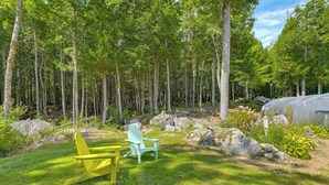 A forested area with a yellow chair and a green Adirondack chair on a grassy lawn. - Explore Coastal Maine! Cozy Apt w/ Yard & Garden (Orland)