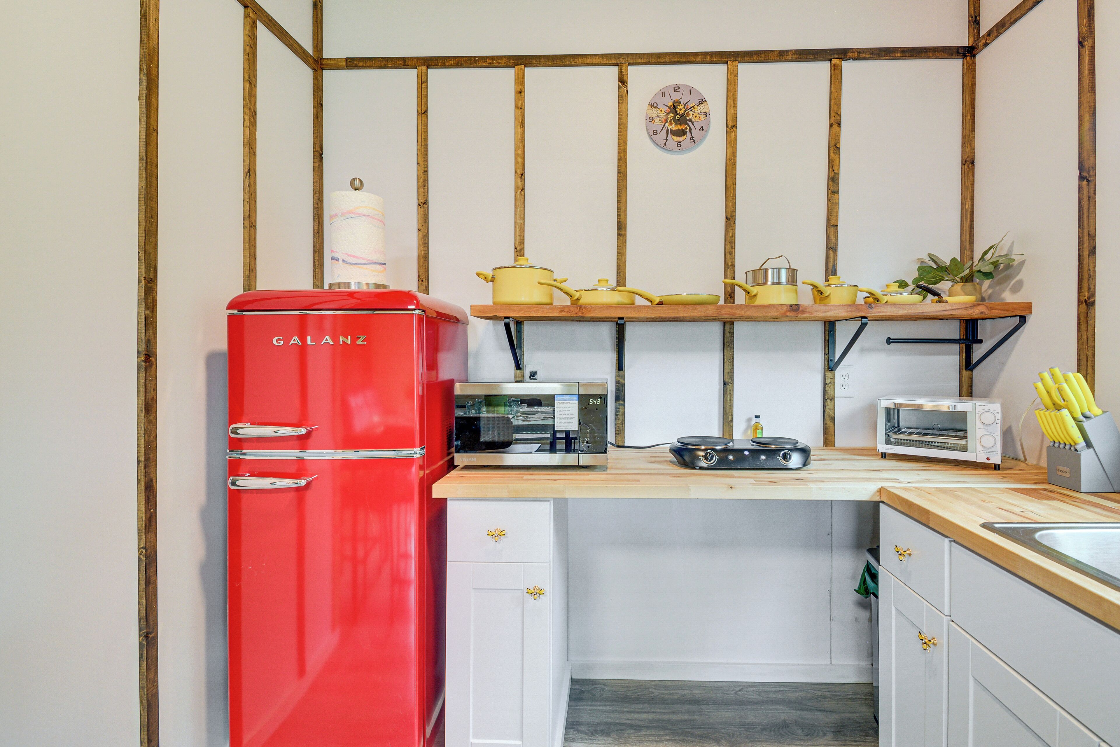 A compact kitchen with a red refrigerator, a wooden countertop, and a wall-mounted shelf with yellow pots.