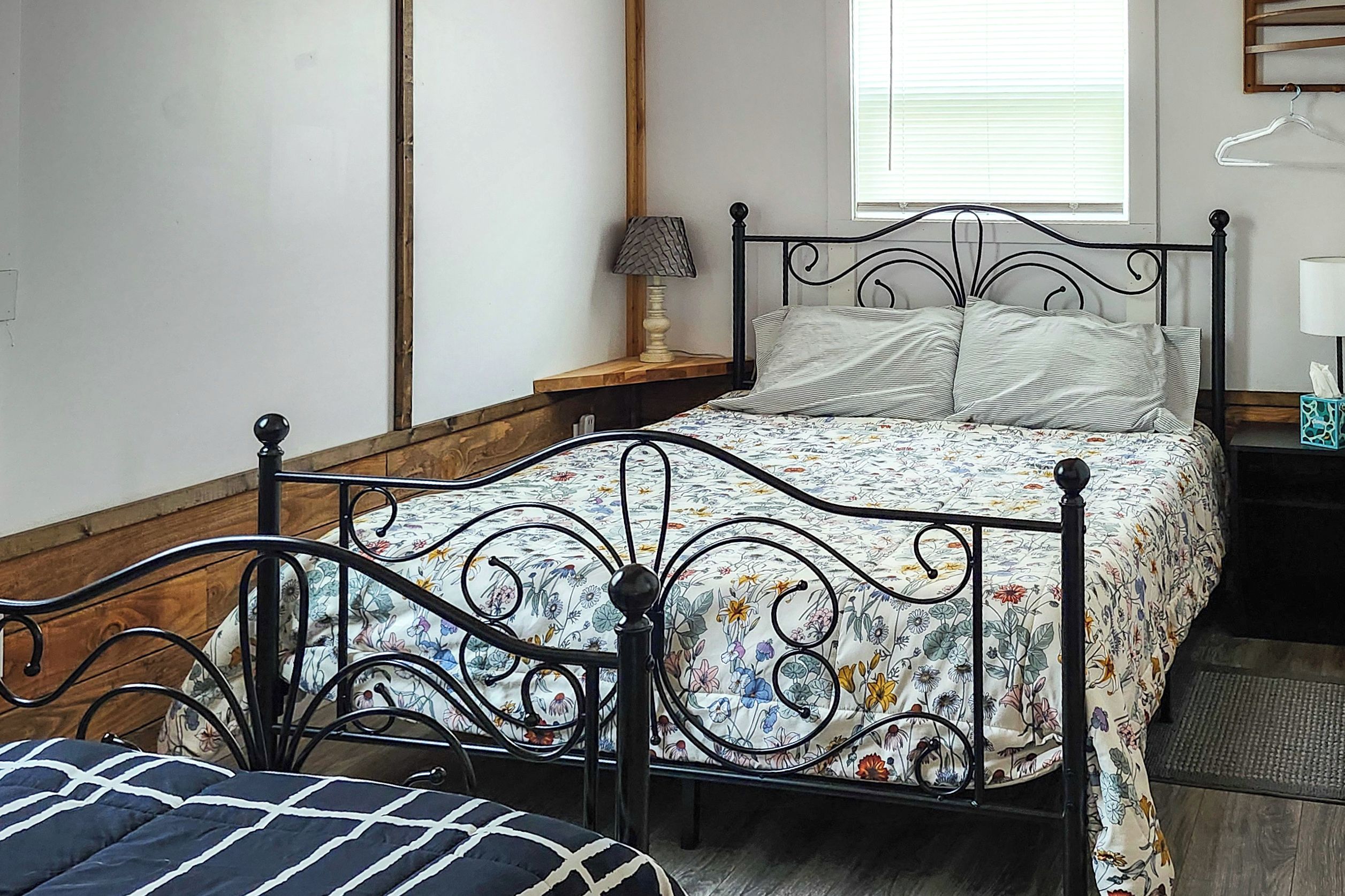 A bedroom with a metal bed frame, a floral bedspread, a bedside table with a lamp, and a window with blinds.