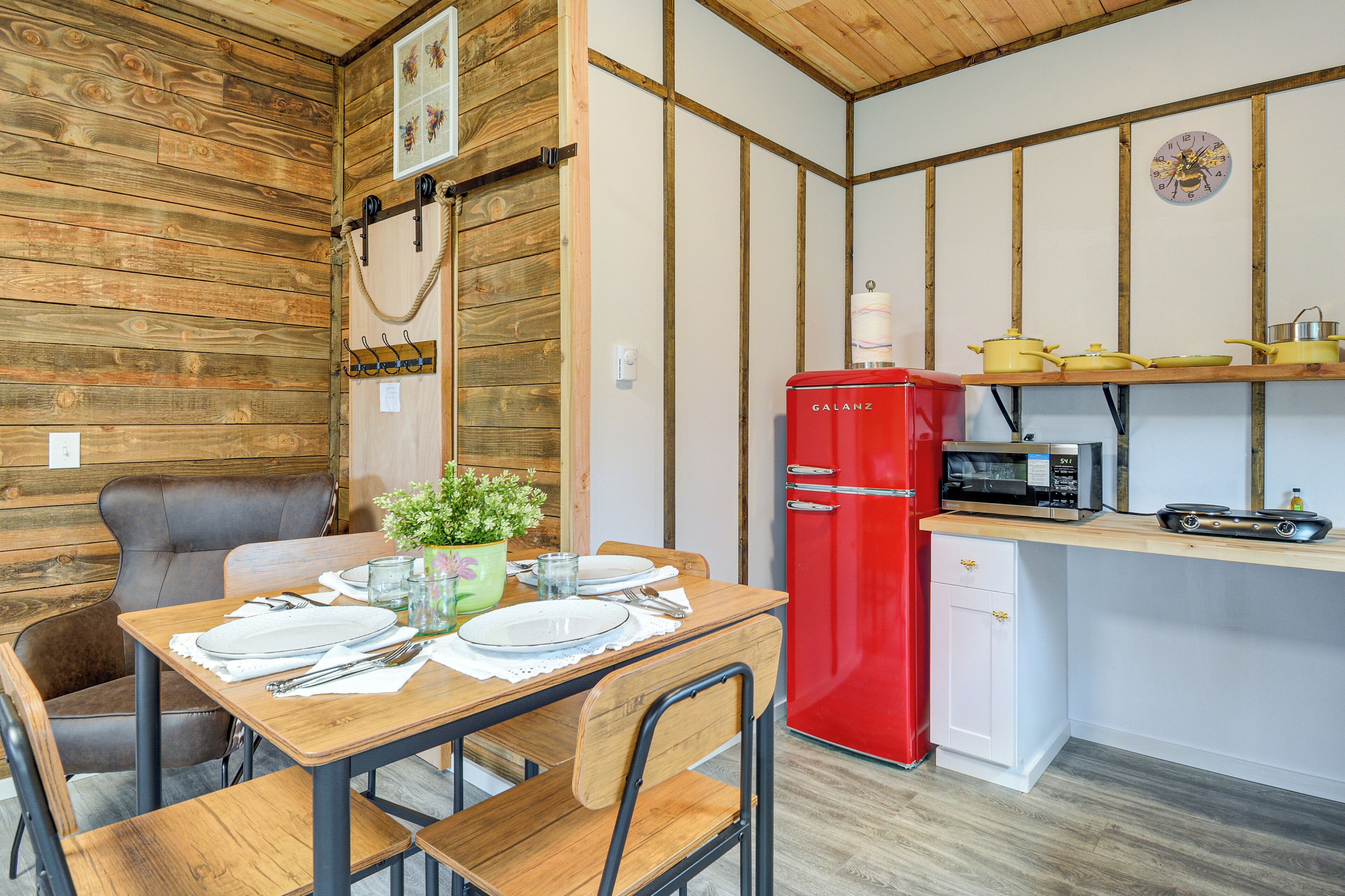 A compact kitchen with a red refrigerator, a microwave, and a small sink. There is a wooden dining table with chairs and a small potted plant.