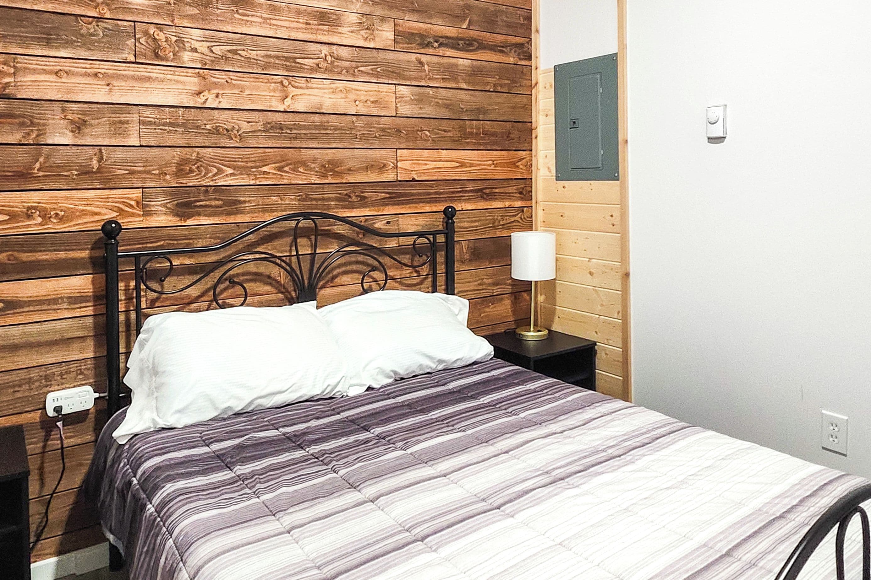 A bed with a headboard against a wooden wall, a bedside lamp, and a power outlet on the nightstand.
